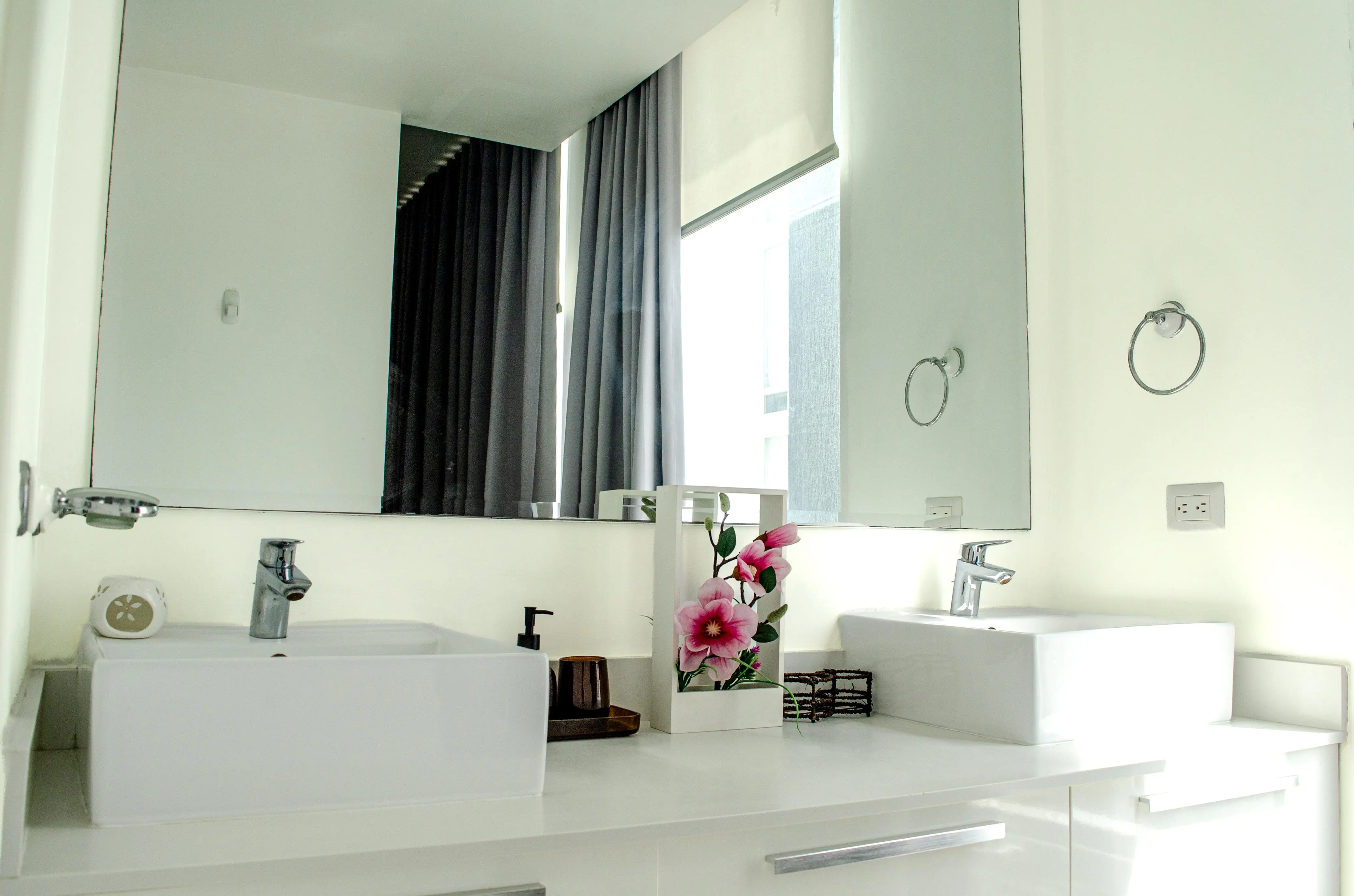 Baño con dos lavamanos blancos, espejo grande y luz vertical junto al espejo.