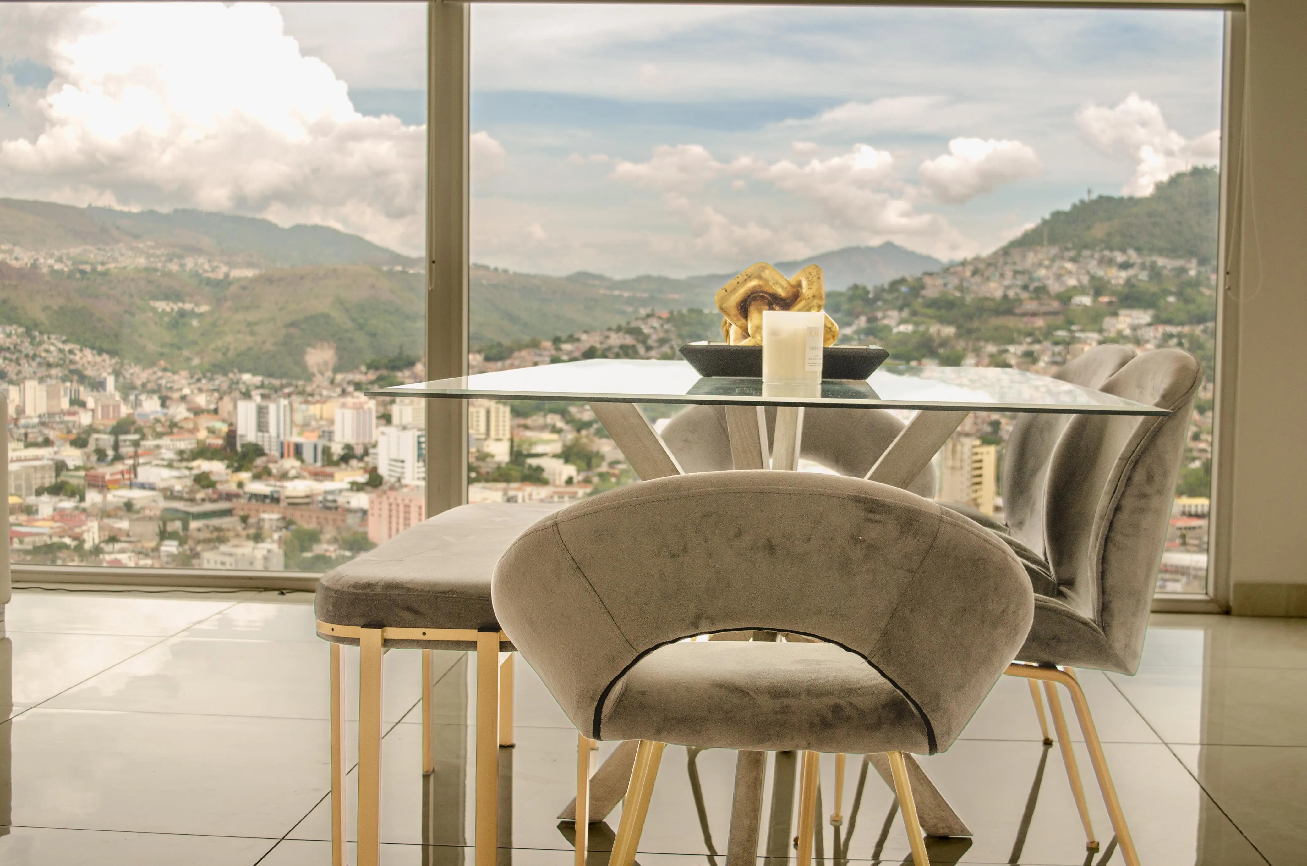Comedor de vidrio con sillas de terciopelo gris frente al ventanal, y la ciudad y las montañas al fondo.
