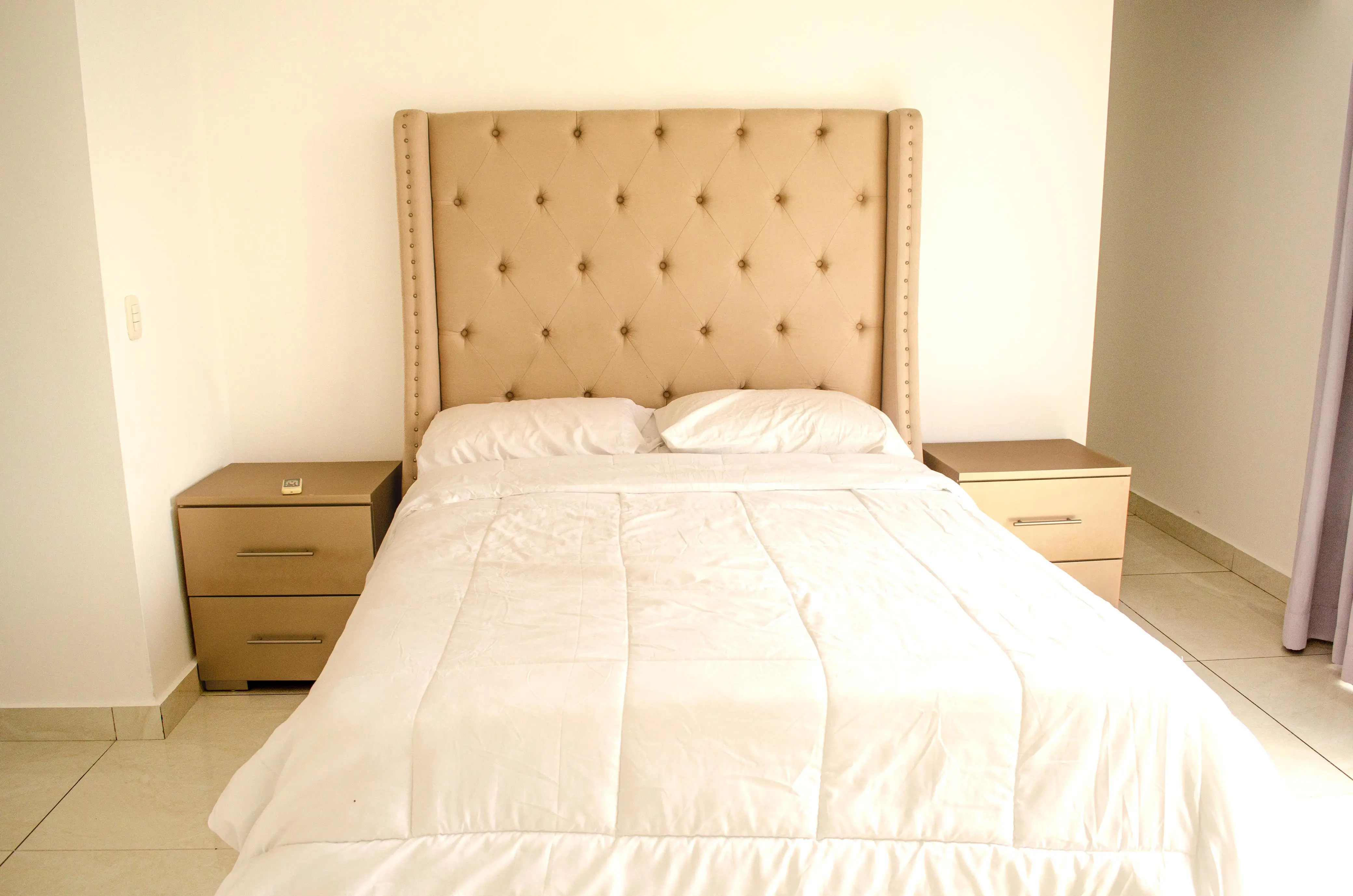 Cama con cabecera capitoné beige, ropa blanca y dos burós.