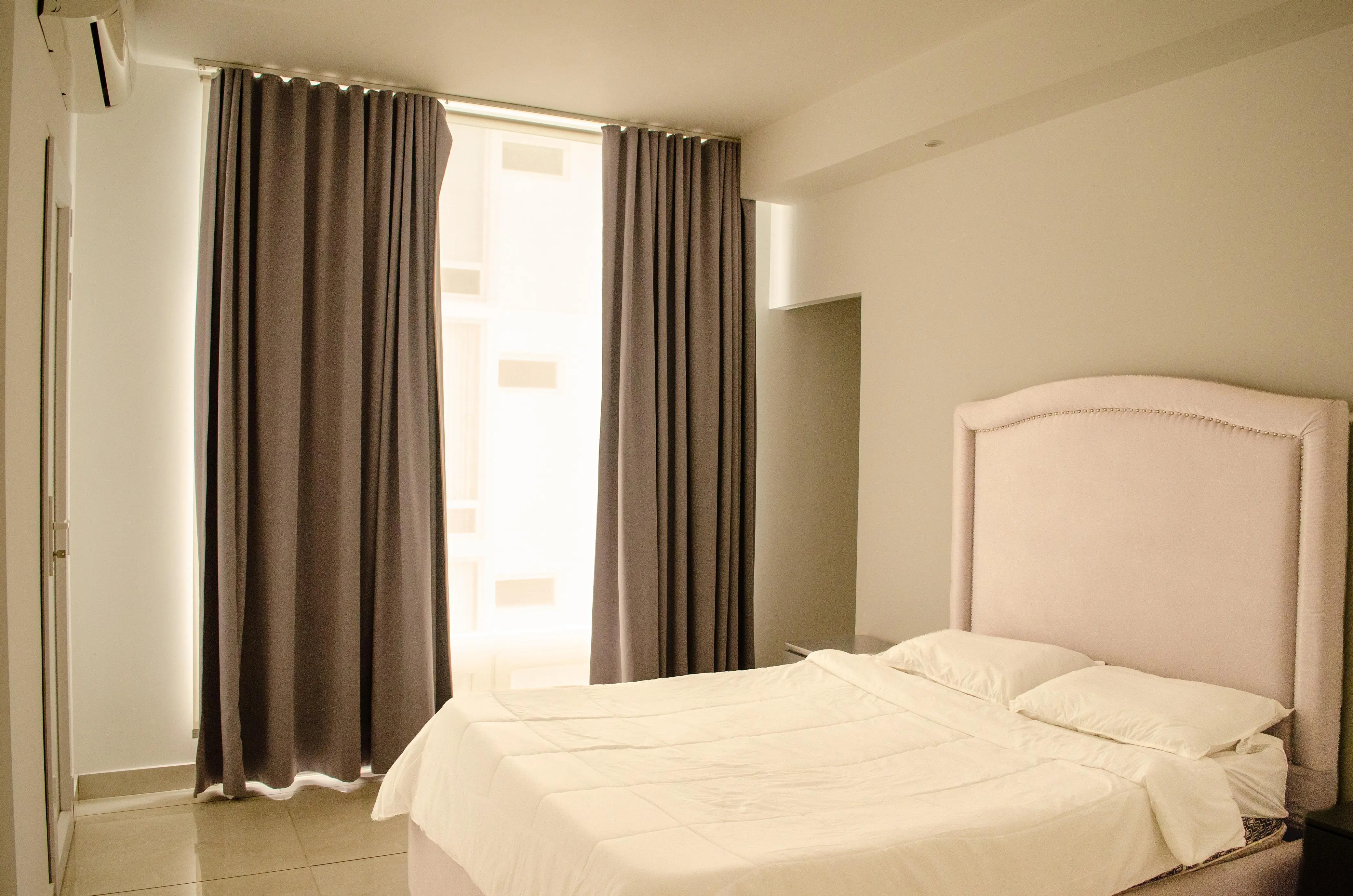 Segunda habitación con cama vestida de blanco, cabecera tapizada y cortinas junto a la ventana.