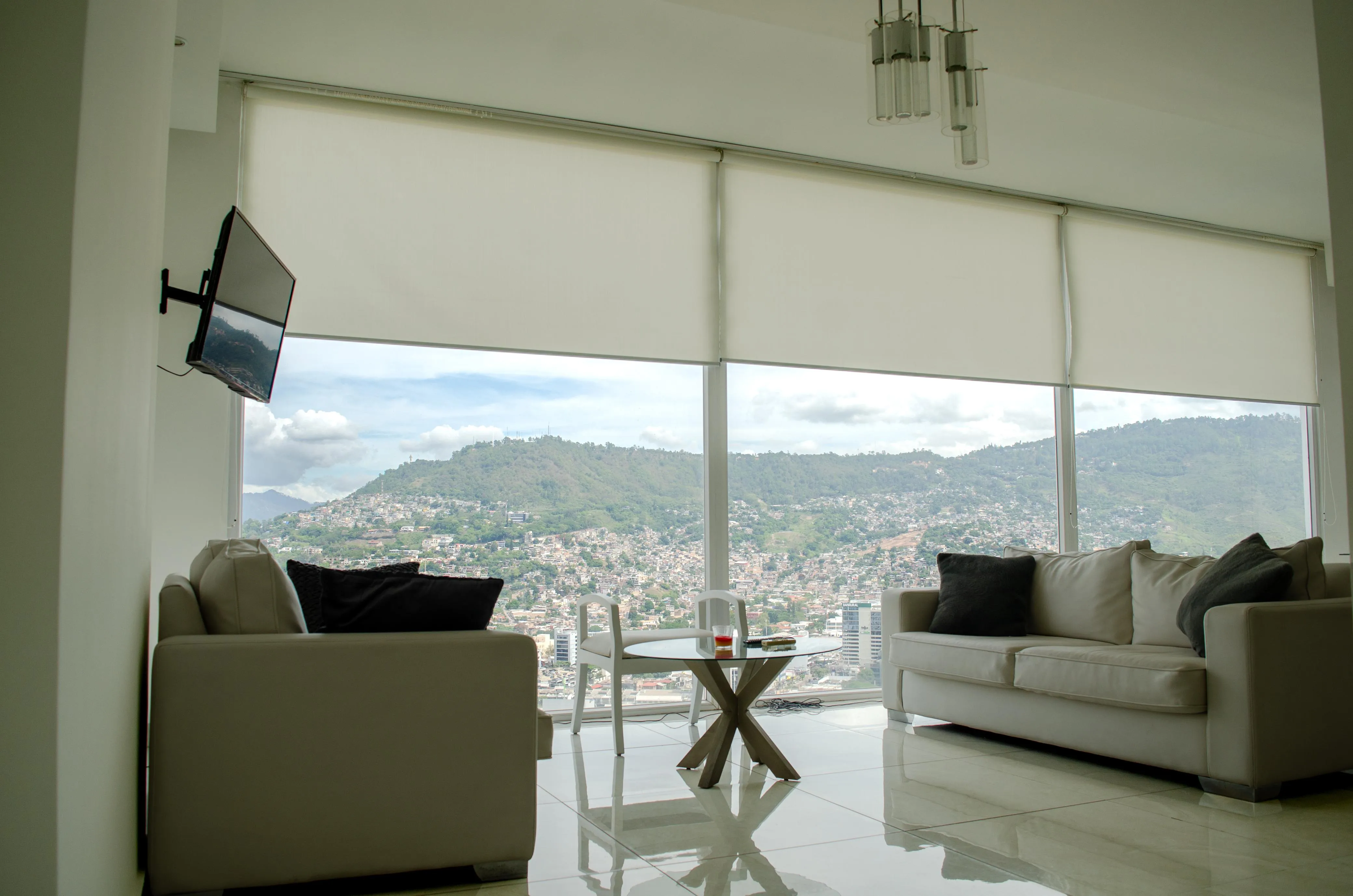 Sala del departamento 1233 con sofá y sillón claros, mesa de vidrio y ventanal con vista a la ciudad y a las montañas.