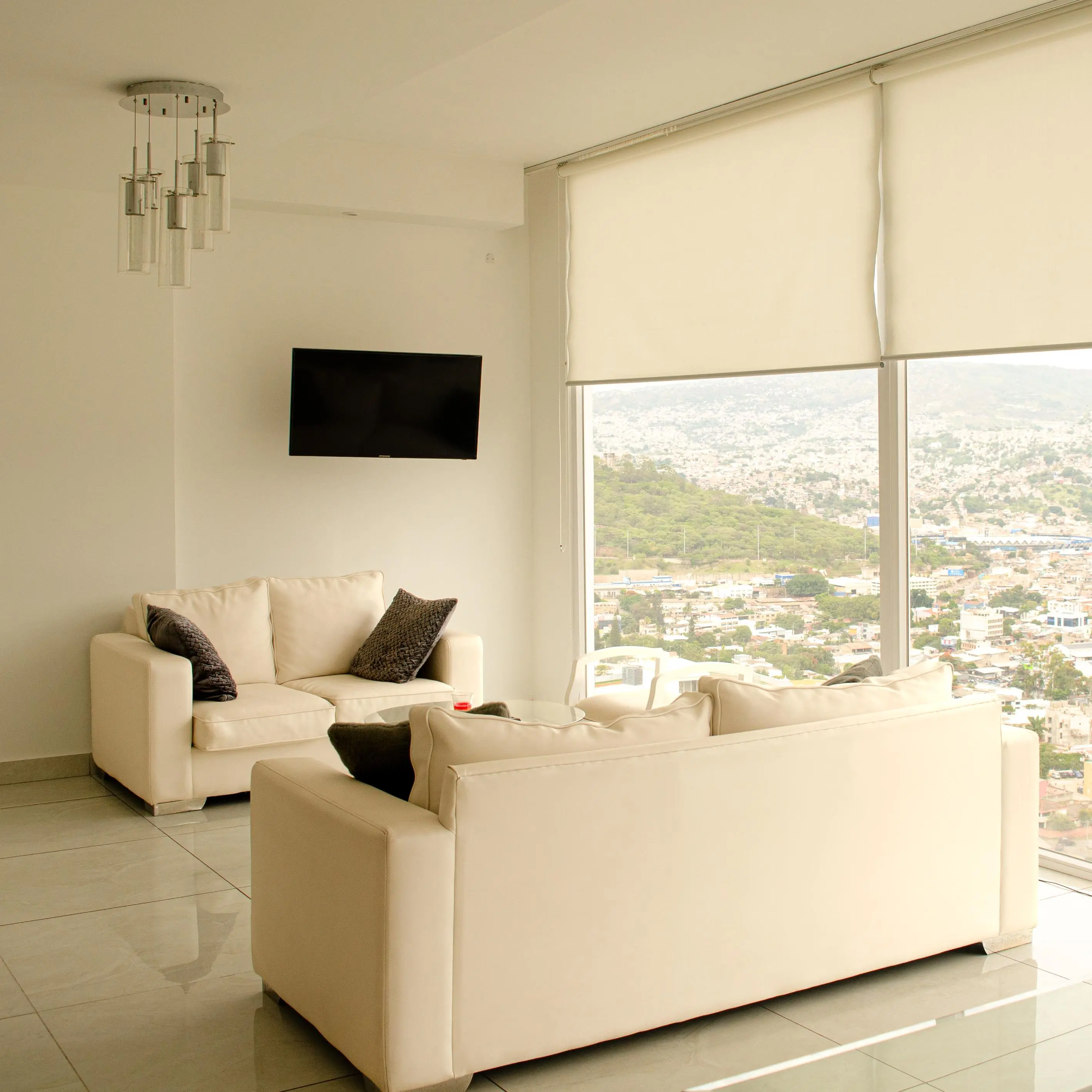 Sala con dos sofás claros, cojines oscuros, televisión en la pared y vista a la ciudad por el ventanal.